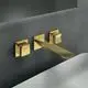 Wand-Waschtischarmatur in Brushed Gold von Ideal Standard mit eckigen Rosetten, zwei Griffen und einem langen Auslauf, montiert an einer dunklen Wand.